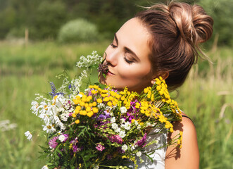 Naklejka premium Summer lifestyle portrait of beautiful young woman smiling and holding bouquet of wild flowers. Standing in the field of grass. Enjoying sun. Happiness and love concept. Сlosed eyes 