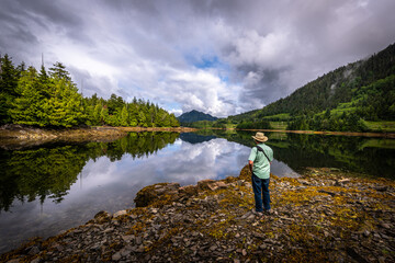 Naklejka na ściany i meble A Man standing at the shore in the morning enjoying the motionless water of the pacific ocean and its mirror like reflection in an inlet near Prince Rupert, British Columbia, Canada.