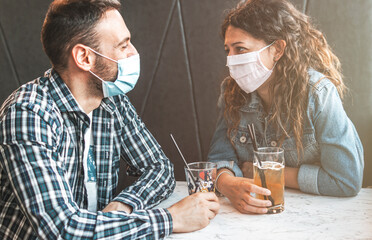 Engaged couple sitting in a caffe bar with surgical masks during the coronavirus pandemic - prevention and social distancing concept.