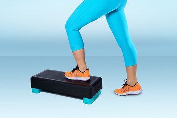 Woman in orange sneakers and turquoise sportswear do exercise on a black-turquoise aerobic and fitness step on blue background