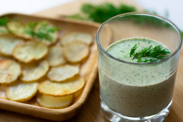 Dill and sour cream dip for homemade potato chips made vegan: healthy snack with dill, tahini, water, vinegar and lemon juice in a glass. Thinly sliced and baked potatoes in the back