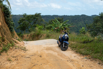 Blue scooter on the dirt road on a hill in tropical jungle with a scenic view on a mountains in a sunny day. Adventure and traveling concept.