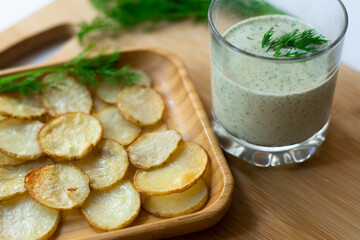 Dill and sour cream dip for homemade potato chips made vegan: healthy snack with dill, tahini, water, vinegar and lemon juice in a glass. Thinly sliced and baked potatoes in the back