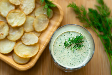 Dill and sour cream dip for homemade potato chips made vegan: healthy snack with dill, tahini, water, vinegar and lemon juice in a glass. Thinly sliced and baked potatoes in the back