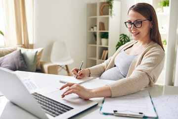 Fototapeta na wymiar Happy young pregnant businesswoman looking at laptop display and making notes