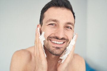 Joyful young man putting shaving soap on his cheeks