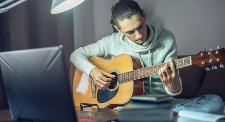 Young male musician is learning to play acoustic guitar in an online lesson using a laptop at night...