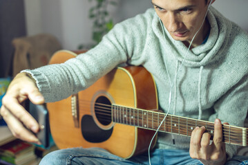 Obraz premium Young musician is learning to play acoustic guitar in an online lesson using phone app. Man is studying courses at home