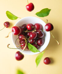 Fresh juicy cherries and leaves fall into a white bowl on a yellow background