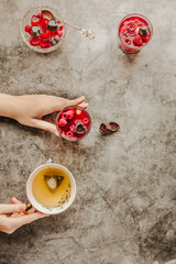 Young woman hands holding and eating jelly dessert with fresh raspberries served in glasses and cup of green tea, on gray background. Top view. Summer or diet food concept