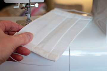 Selective focus on white handmade fabric cotton face mask on sewing machine. Woman sew face mask at home. Protection against saliva, cough, dust, pollution, virus, bacteria, COVID-19, coronavirus.