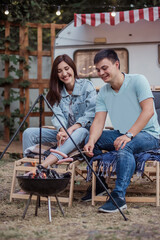 Happy young family at a campsite fries sausages on a bonfire. Picnic of a beautiful couple in love at the trailer, sitting comfortably, hugging, battle on sausages, kissing, laughing. Rest in nature