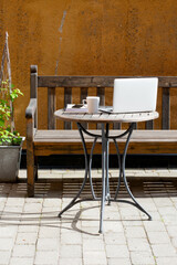 Concept of working from home outdoors. Laptop, cup of coffee, notebook and mobile phone on wooden cafe table. Wooden bench or sofa with a green plant next to it, in front of a orange building wall.