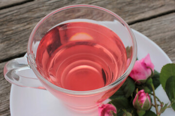 Tea rose herbal tea in a glass cup with pink buds and flowers on a rustic wooden table. Summer tea party outdoors
