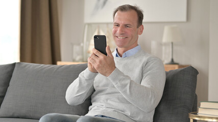 Middle Aged Businessman Celebrating Success on Smartphone on Sofa