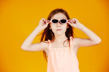 jolie jeune enfant fille caucasienne aux cheveux longs châtains portant des lunettes solaires en souriant sur le thème des vacances d'été sur fond jaune de studio - obrazy, fototapety, plakaty