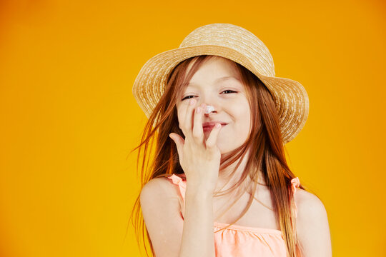 jolie jeune enfant fille caucasienne aux cheveux longs châtains mettant de la crème solaire sur son visage pour protéger sa peau sur fond jaune de studio
