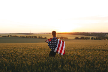 Beautiful girl with the American flag in a wheat field at sunset. 4th of July.  Independence Day.