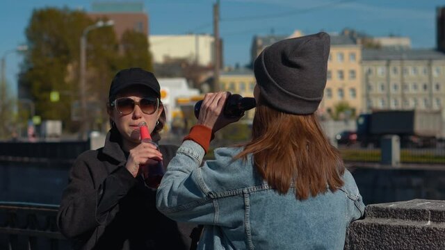 Two young girls drink alcohol from glass jars on the street. Female alcoholism in a public place. Drinking beer for the sake of meeting and good mood.