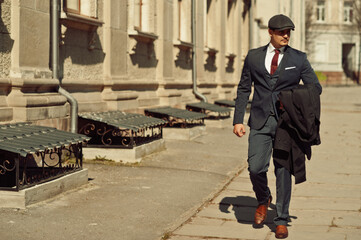 Portrait of retro 1920s english arabian business man wearing dark suit, tie and flat cap walking on old streets.