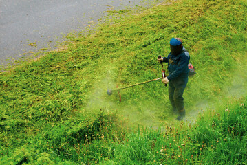 A man mows grass with a gasoline scythe in a protective uniform