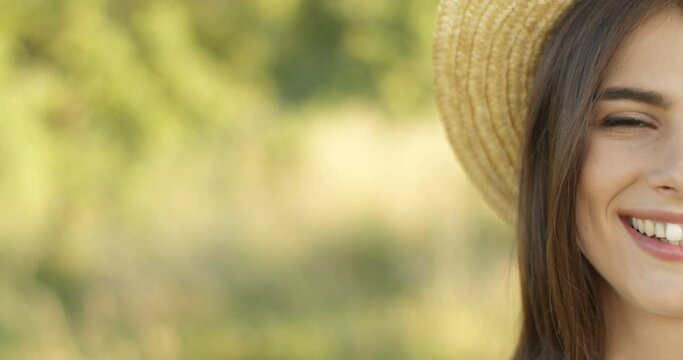 Close up of half face of pretty joyful Caucasian brunette girl in hat smiling happilly to camera outdoor in summer. Portrait of charming young female in good mood with smile at nature.