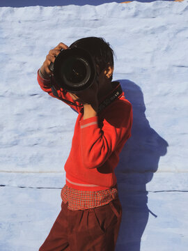 Young teenager photographer boy aiming with a camera and big lens in front of his face. Blue background and wearing an orange sweater.