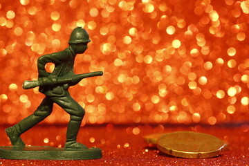 War and money concept. Close up of toy soldier and coins on a blurred red sparkling background....