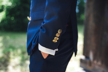 Side view of man in blue suit with hand in pocket. Close up of jacket sleeve gold color buttons with anchor sign. Outdoor background.