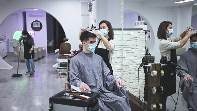 Young woman stylist wearing face mask and gloves working in hairsalon doing haircut and hair styling for male client, new normal due to coronavirus outbreak