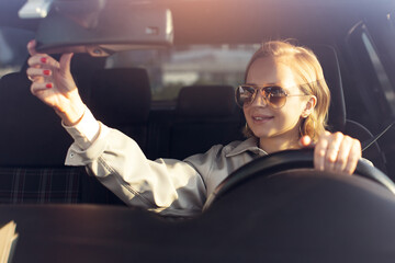 Woman in sunglasses, new driver sitting in car, riding on road, adjustment of mirror. Holding steering wheel. Riding on vacation with sunlight, enjoyment and relax