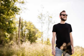 Naklejka na ściany i meble A young unshaven man in his 30s wearing dark shades and a black, round-neck shirt smiling, confident man with facial hair wearing dark shades blurred background.