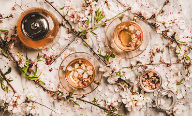 Flat-lay black tea with dry rose flowers in cups and teapot over white marble background with spring bloossom almond tree branches, top view