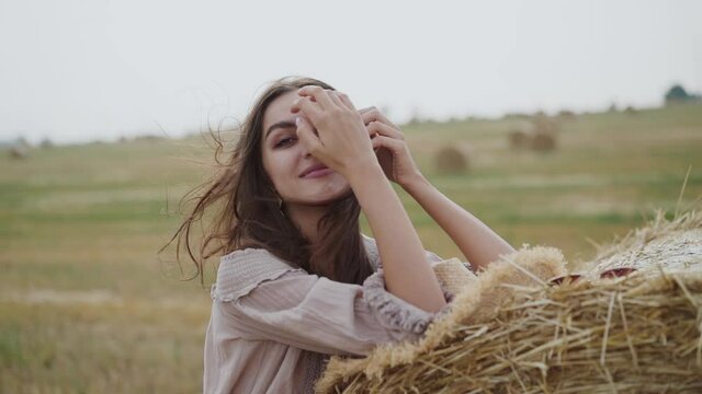 Happy girl leaned on haystack in a field, correcting blowing hair and smiling