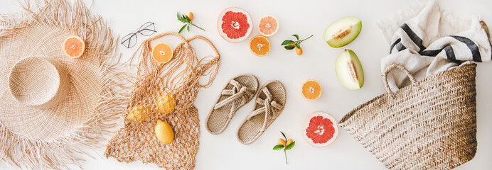 Summer mood layout. Flat-lay of summer natural sandals, straw sunhat, beach rafia and net bag, striped beach towel, sunglasses and fresh fruits over white plain background, top view
