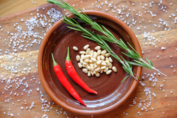 Fresh rosemary, hot red pepper and pine nuts on a clay plate on a wooden background with crystals of coarse white salt. The concept of a delicious and healthy diet.