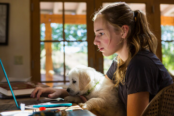 teen age girl holding her English Cream Golden Retriever puppy as she types on her laptop computer