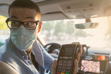 Taxi driver wearing glasses and mask to protect himself from coronavirus hands a payment terminal to a customer inside his car