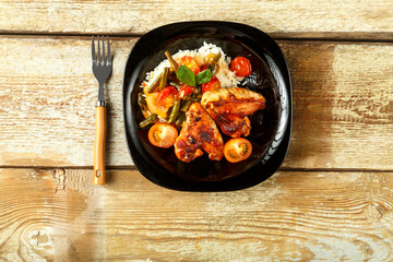 Baked chicken wings in tomato sauce with rice and vegetables on a black plate on a wooden table.