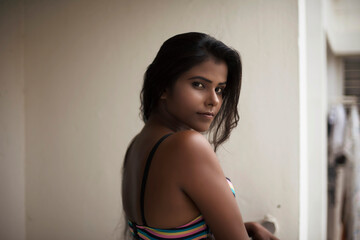 Portrait of an young and beautiful dark skinned Indian Bengali woman in colorful lingerie / bikini and hot pants posing in casual mood on a balcony in white urban background. Boudoir photography.