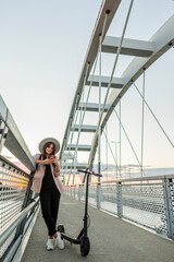 A young girl is leaning on the bridge fence while typing on her phone. An electric scooter is parked next to her.