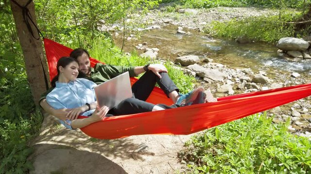 Loving couple lies in a hammock and watches a movie on a laptop. Spending good time outdoor.