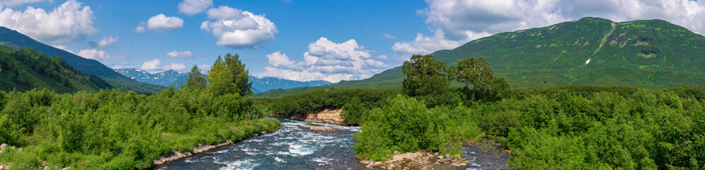 Naklejka na ściany i meble Panorama view of beautiful summer landscape - stream water of mountain river and green forest on hills along riverbank on sunny day with white clouds in blue sky. Headline summertime environment.