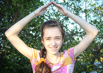 A smiling teenage Girl makes a roof over her head with her hands against the background of trees.