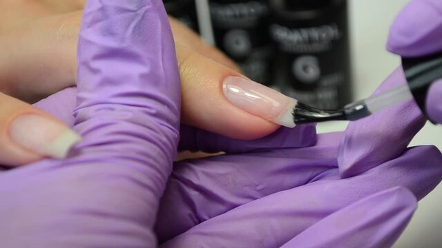Manicure master in purple gloves apply transparent nail Polish on the client's nail.