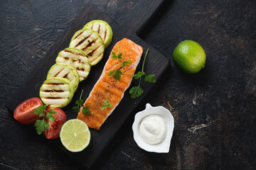 Roasted salmon fillet with zucchini slices and tomatoes, above view on a dark brown stone background with space, horizontal shot