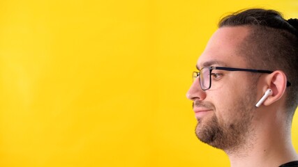 Obraz premium Displeased person man with glasses and headphones looks to the left side of the screen. Profile portrait of an attractive adult unshaven man on a yellow background. Upset emotional guy keeps calm