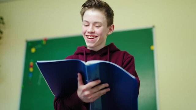 Smiling schoolboy with a folder. Teenage boy standing in the classroom and looking into the school register. Portrait of a happy teenager in the school.