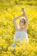 beautiful blonde girl with long hair in a rapeseed field in a white dress in a field of yellow flowers blooming in a rapeseed field at sunset or sunrise
