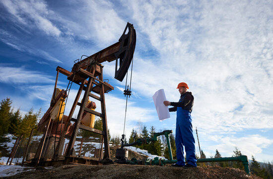 Full Length Of Oil Man In Work Overalls And Helmet Holding Sheet Of Paper While Standing Near Oil Pumping Unit. Male Worker Studying Plan Of Oil Field, Checking Work Of Pump Jack Under Beautiful Sky.